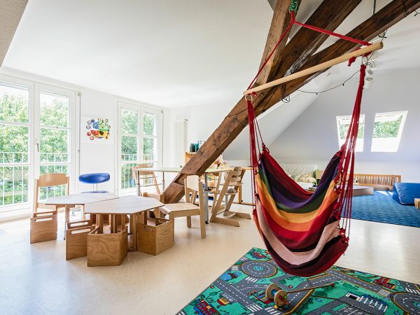 Das Bild zeigt einen Gruppenraum in unserem Krippenhaus. Vorne hängt ein bunter Schaukelstuhl, auf dem Boden liegt ein Verkehrsteppich. Links im Bild sind Tische und Stühle. Der Raum hat große Fenster und ist sehr hell.