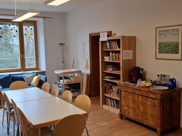 Das Bild zeigt unser Familiencafé mit einem Besprechungstisch, einer Kommode, einem Bücherregal, einem Sofa und einem Wickeltisch.