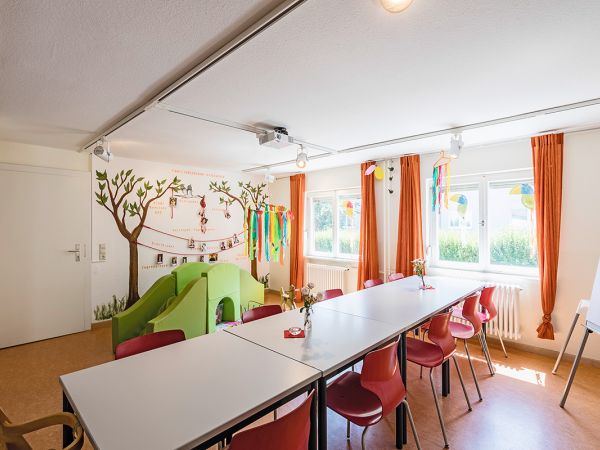 Das Familiencafé im Familienzentrum Stockacker mit einem langen Tisch, orange Vorhängen, einer grünen Kinderrutsche und einem Flipchart.