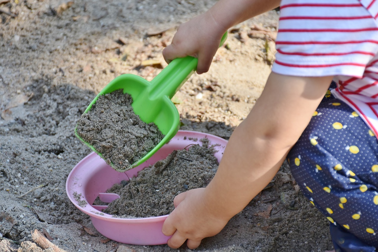 Ein kleines Kind spielt im Sandkasten mit einer grünen Schaufel und einem rosa Sieb.